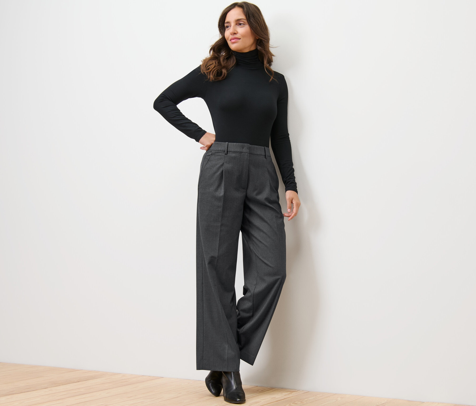 Kobieta pozuje oparta o ścianę w czarnym golfie i wygodnych spodniach z tkaniny.