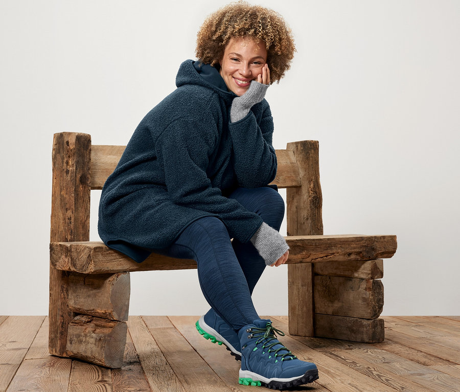 Kobieta siedzi na drewnianej ławce, ubrana w pluszowy płaszcz oversize i legginsy sportowe.