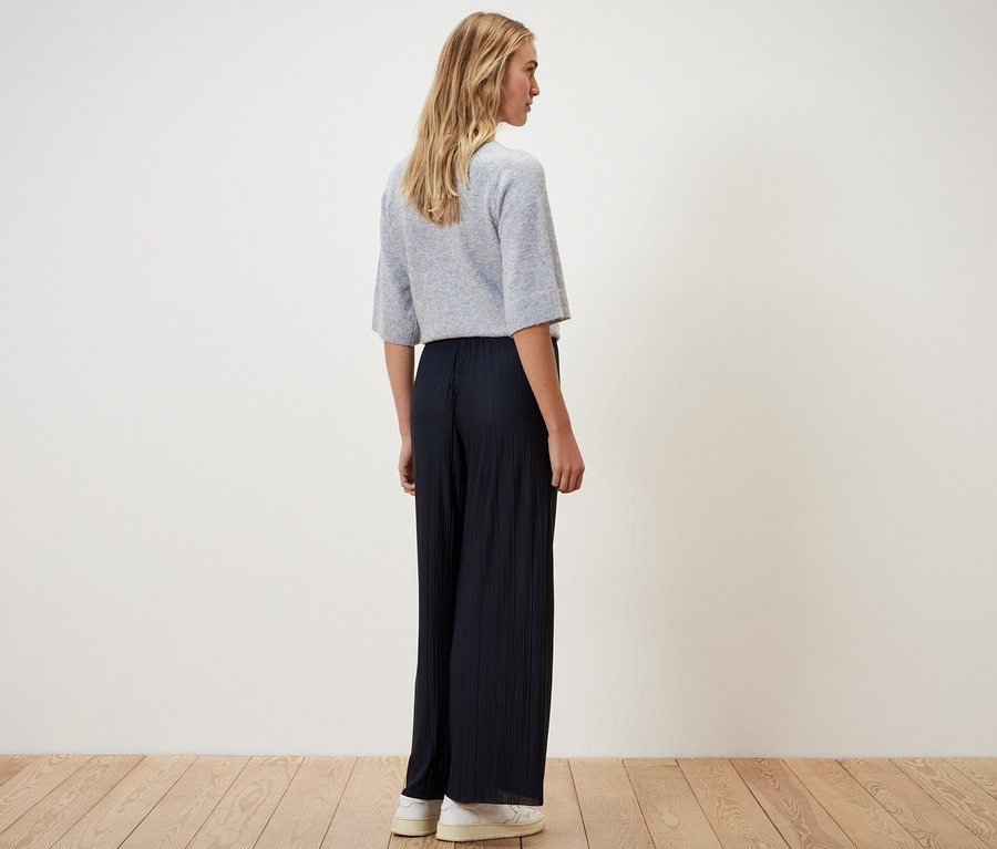 Kobieta stoi tyłem do kamery w granatowych plisowanych spodniach.