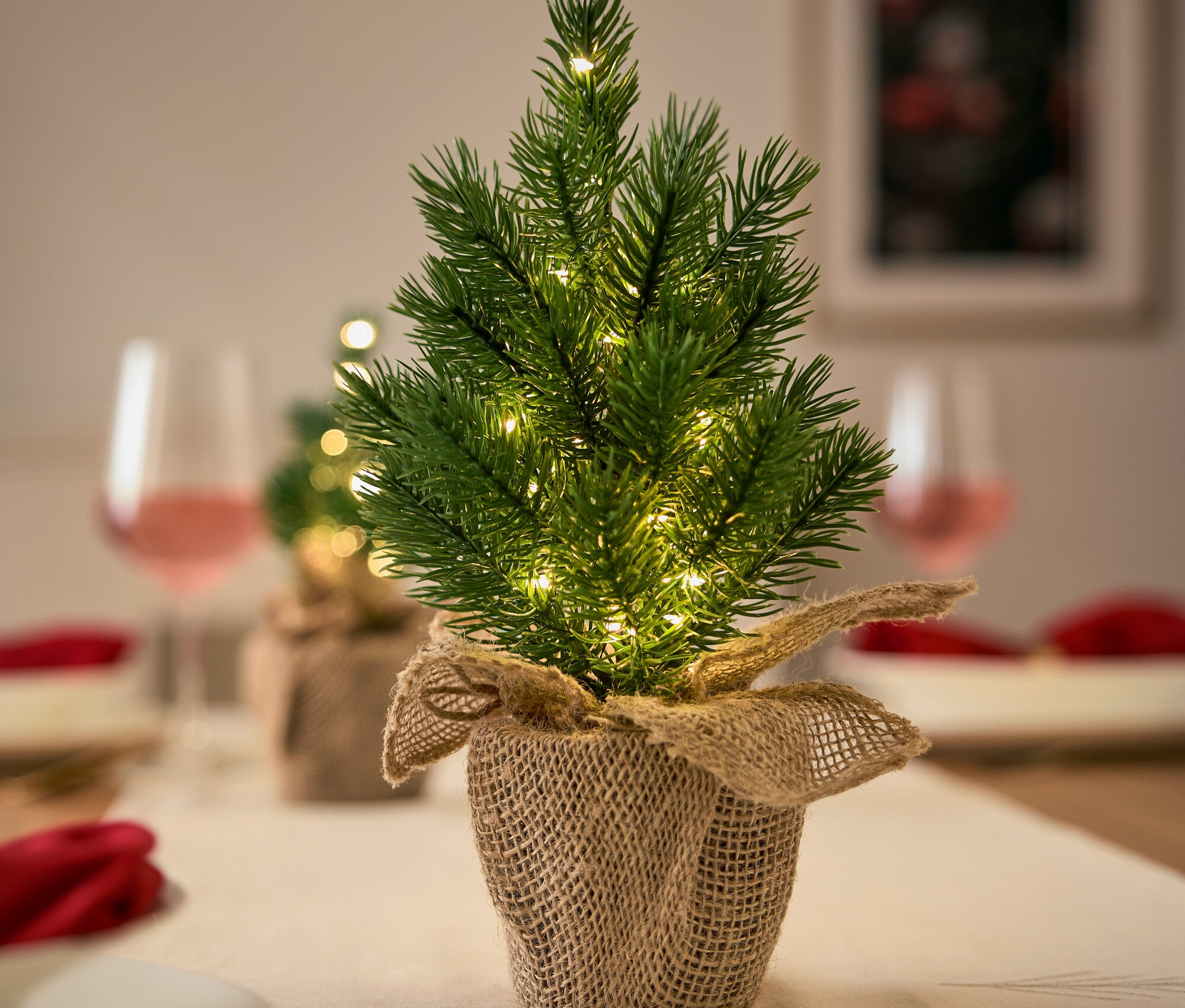 Mała choinka z lampkami w jutowej doniczce na świątecznie nakrytym stole.