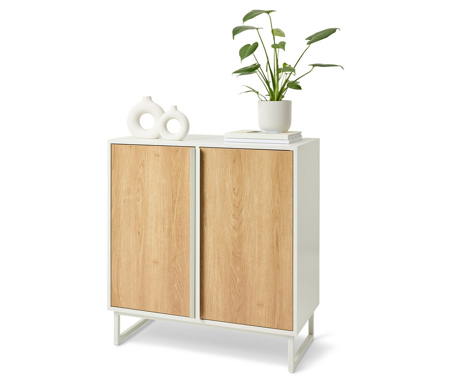 Biała szafka z drewnianymi drzwiami. Na górze szafki znajdują się dwa białe wazony, książka i roślina w białej doniczce.