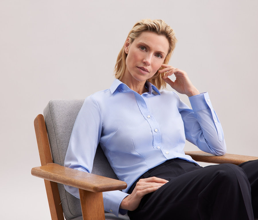 Blondynka siedzi w szarym fotelu, ubrana w błękitną koszulę i czarne spodnie.