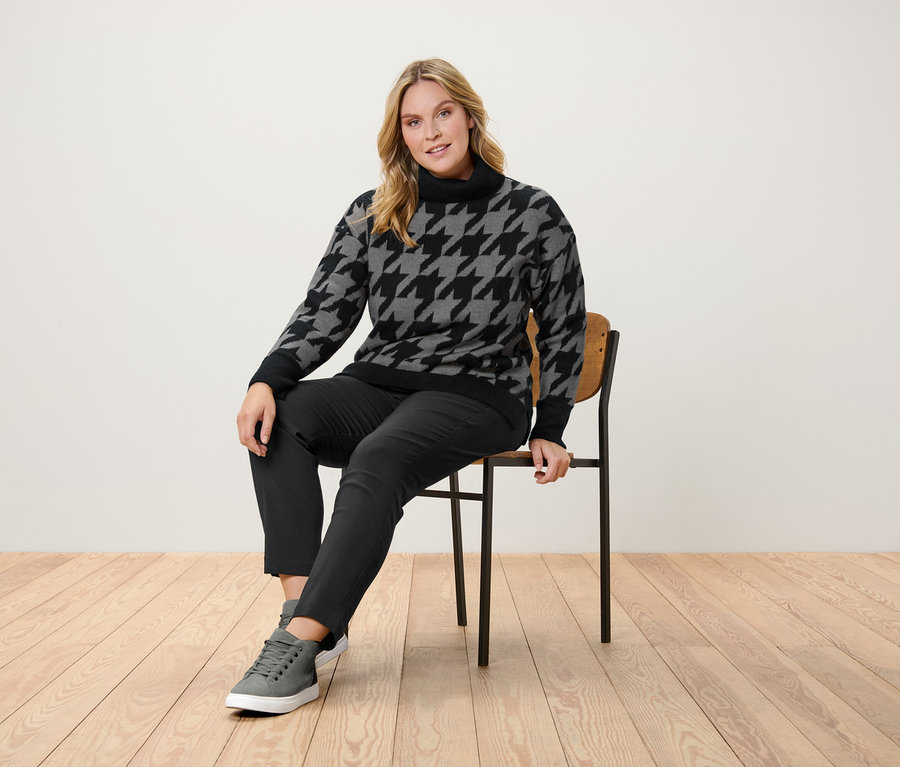 Kobieta siedzi na krześle ubrana w sweter w pepitkę, czarne spodnie i szare tenisówki.