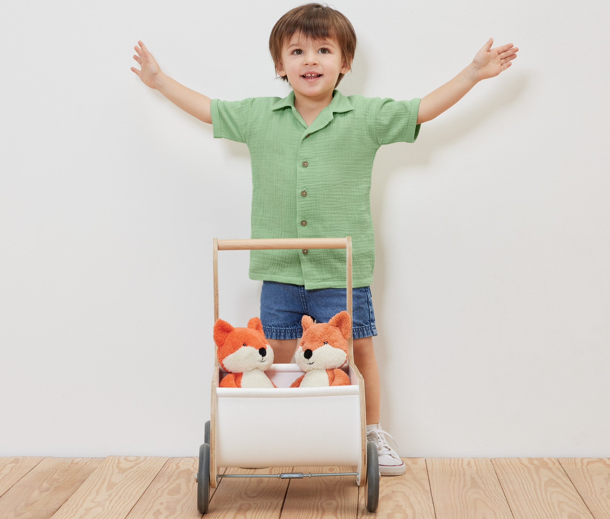 Chłopiec stoi z rozłożonymi ramionami za wózkiem z dwoma pluszowymi lisami, ubrany w muślinową koszulę z kołnierzykiem.