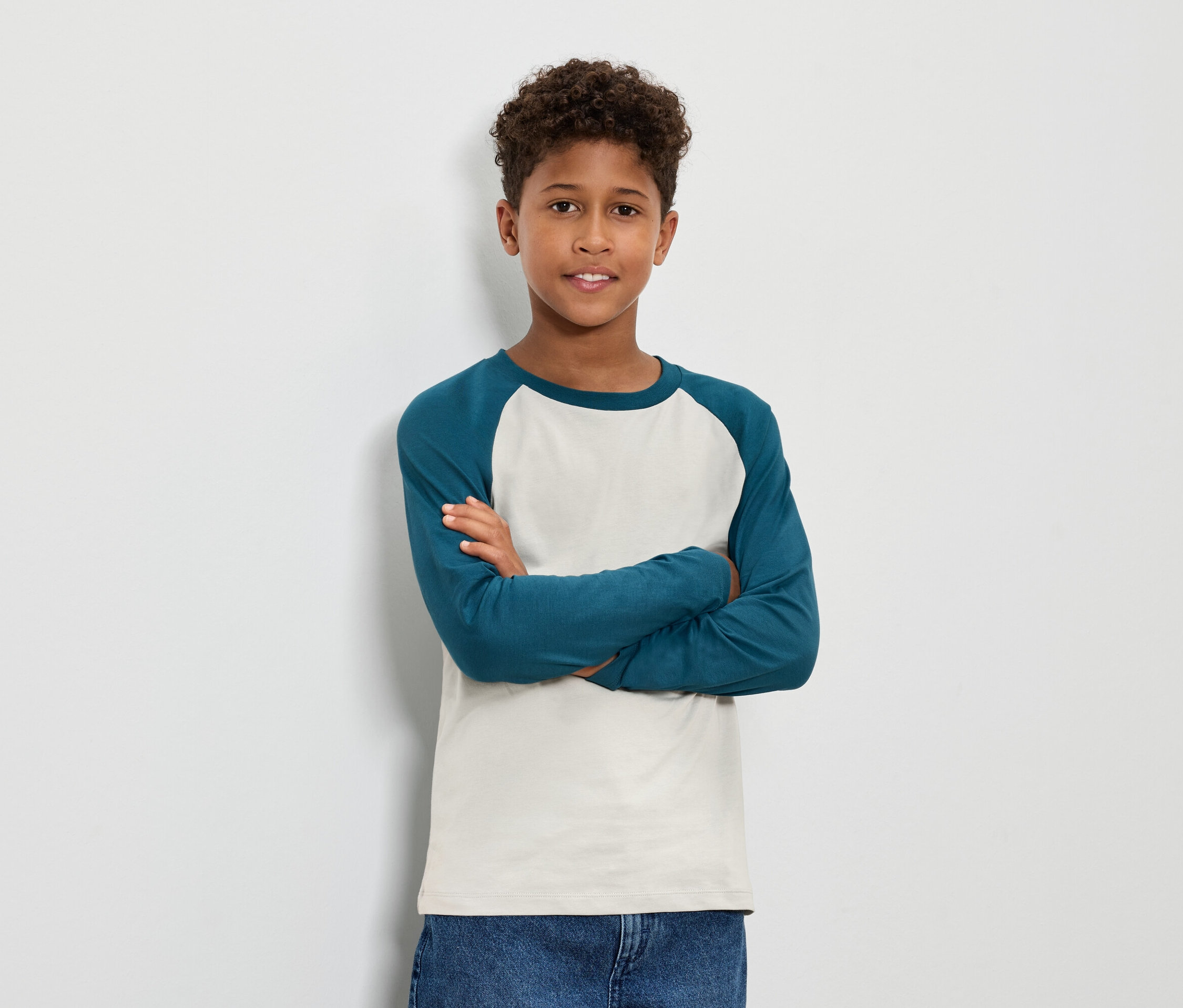 Chłopiec z brązowymi kręconymi włosami stoi ze skrzyżowanymi ramionami, ubrany w niebiesko-zieloną koszulkę z długim rękawem i dżinsy.