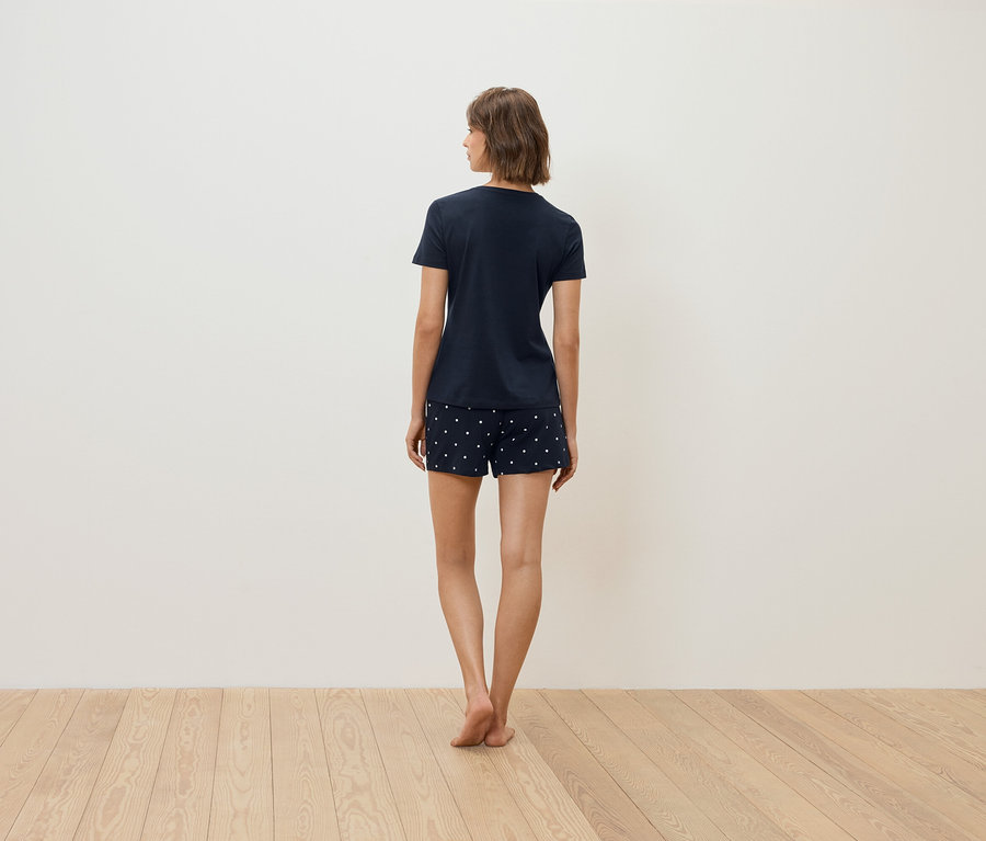 Kobieta stoi tyłem do aparatu, ubrana w granatowy T-shirt i szorty w białe kropki.