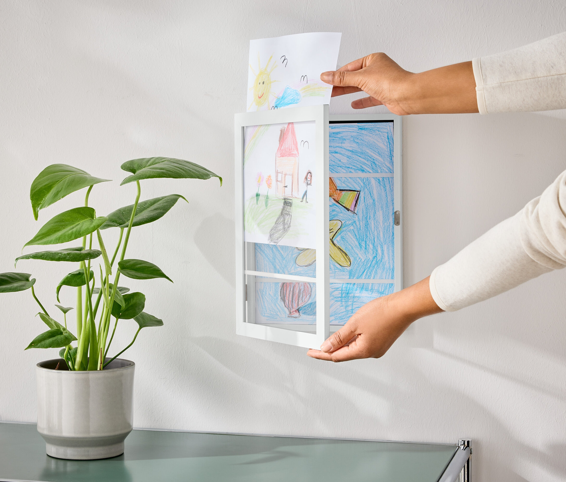 Ręce wkładają rysunek w rozkładaną ramkę na zdjęcia, która wisi na ścianie obok rośliny doniczkowej.