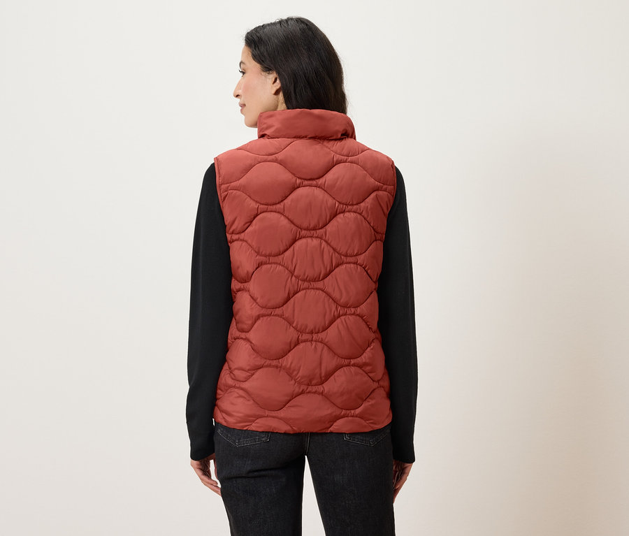 Kobieta tyłem do kamery ma na sobie pomarańczową pikowaną kamizelkę i czarną koszulę z długim rękawem.