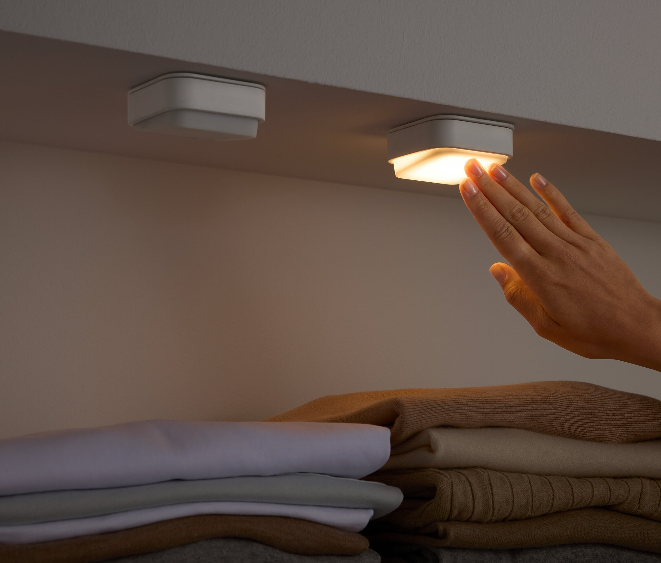 Ręka dotyka kwadratowego światła pod półką nad złożonymi ubraniami.