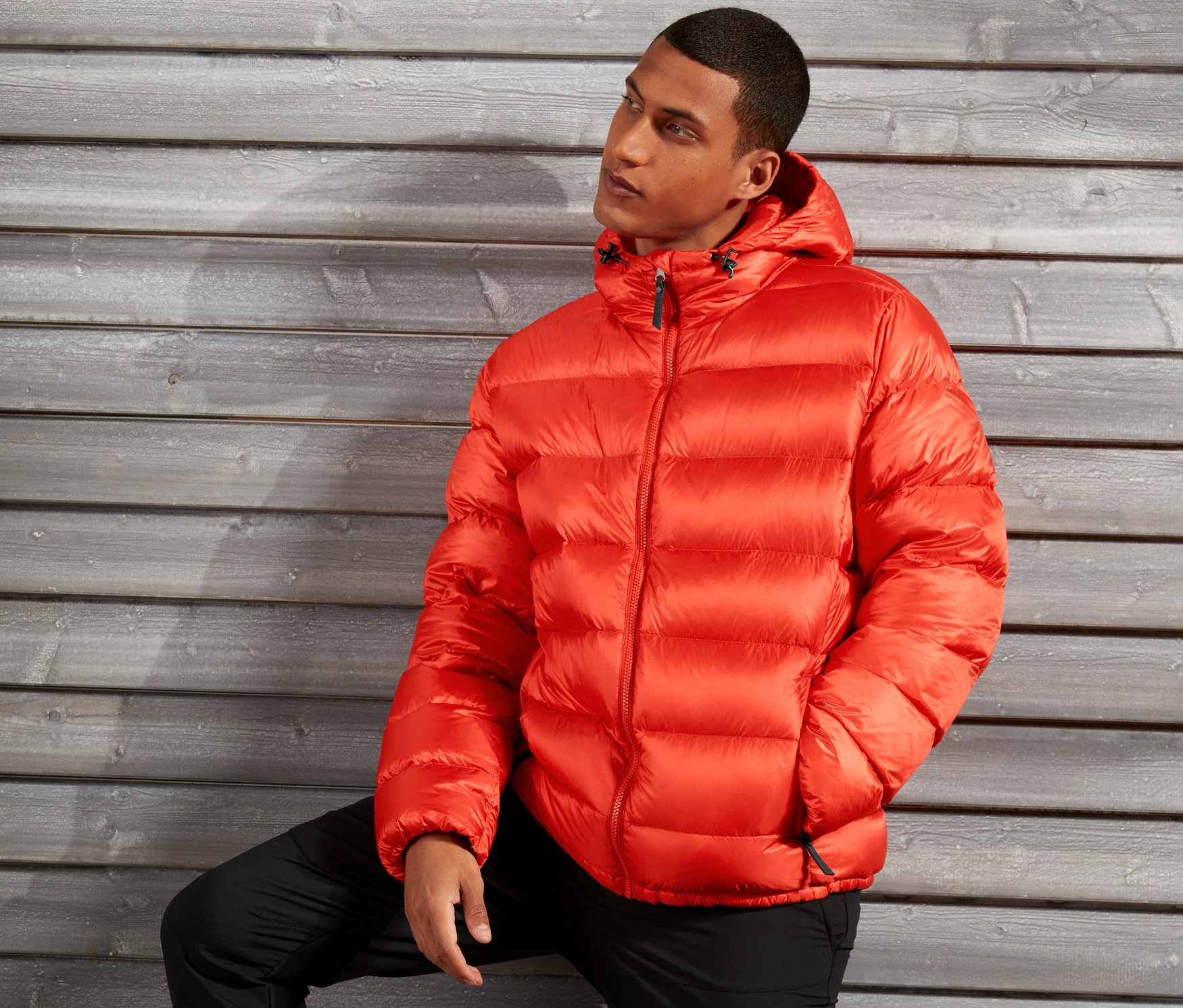 Mężczyzna pozuje w pomarańczowej kurtce puchowej na tle drewnianej ściany.