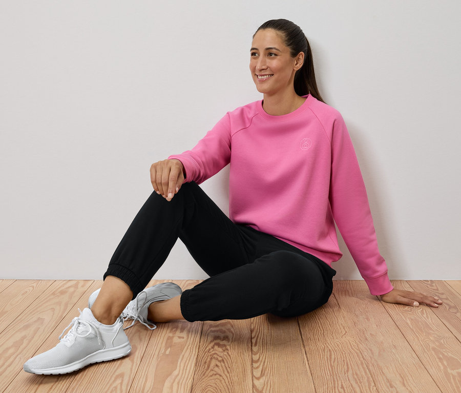Kobieta siedzi na drewnianej podłodze, ubrana w różową bluzę, czarne spodnie i białe tenisówki.