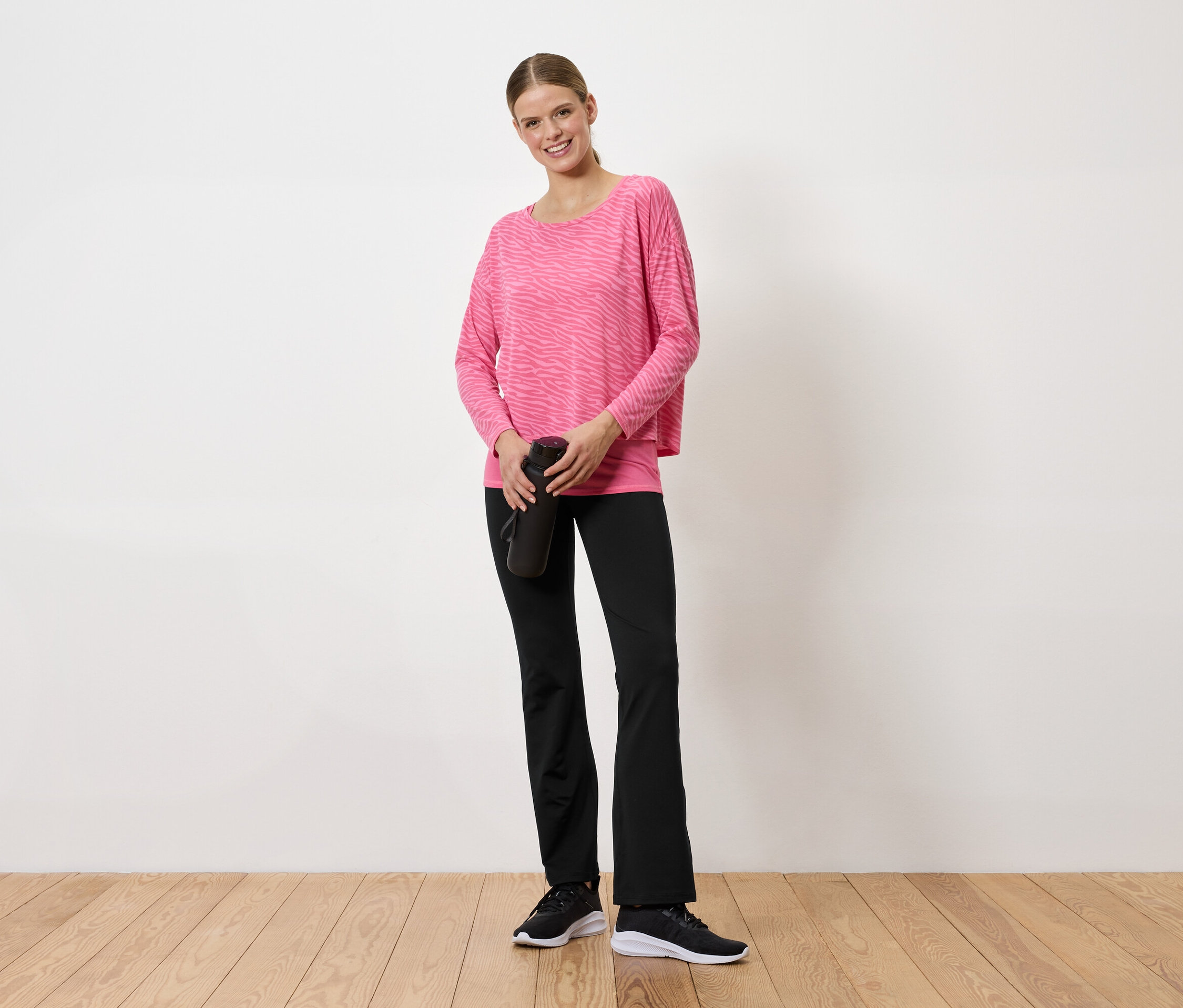 Kobieta w różowym swetrze i czarnych spodniach trzyma w rękach czarną butelkę na wodę.