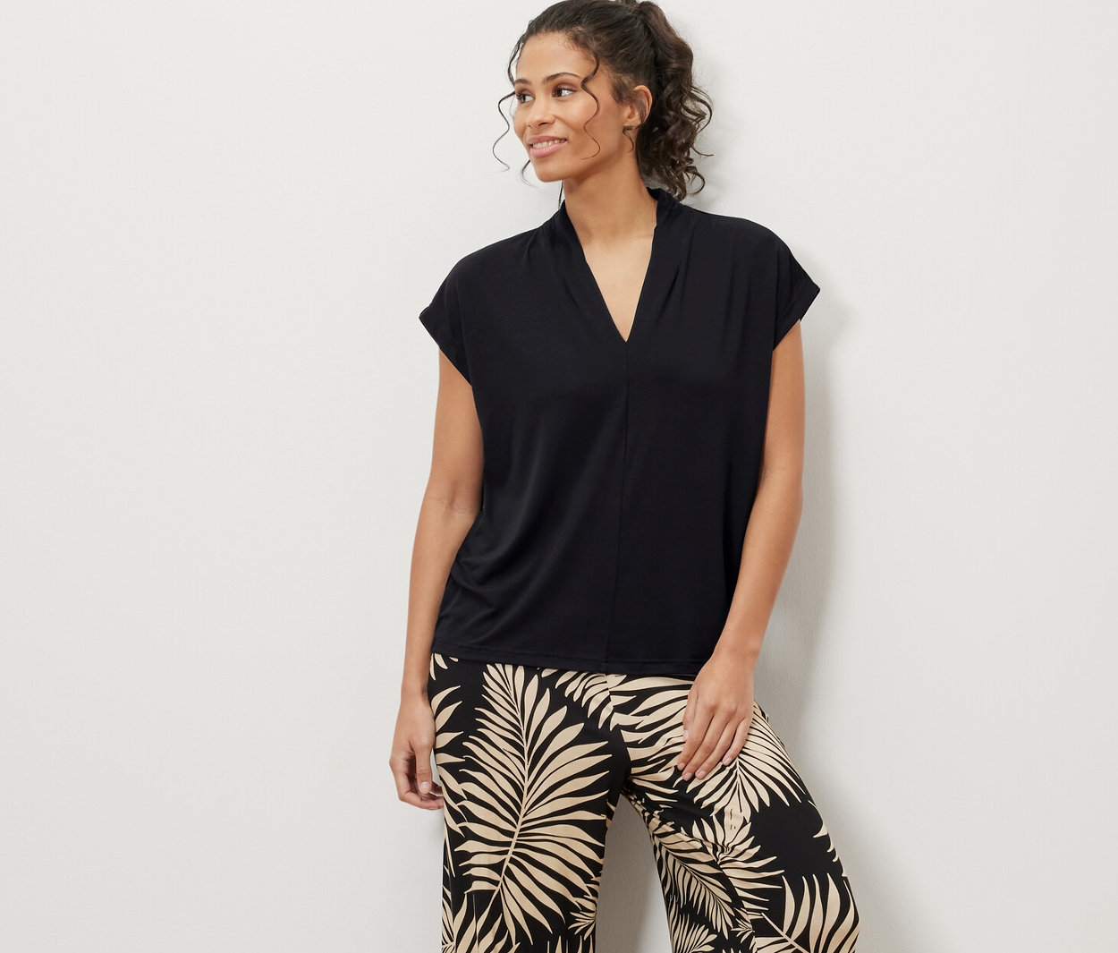 Kobieta stoi i ma na sobie czarną bluzkę z dekoltem w serek i spodnie we wzór liści.