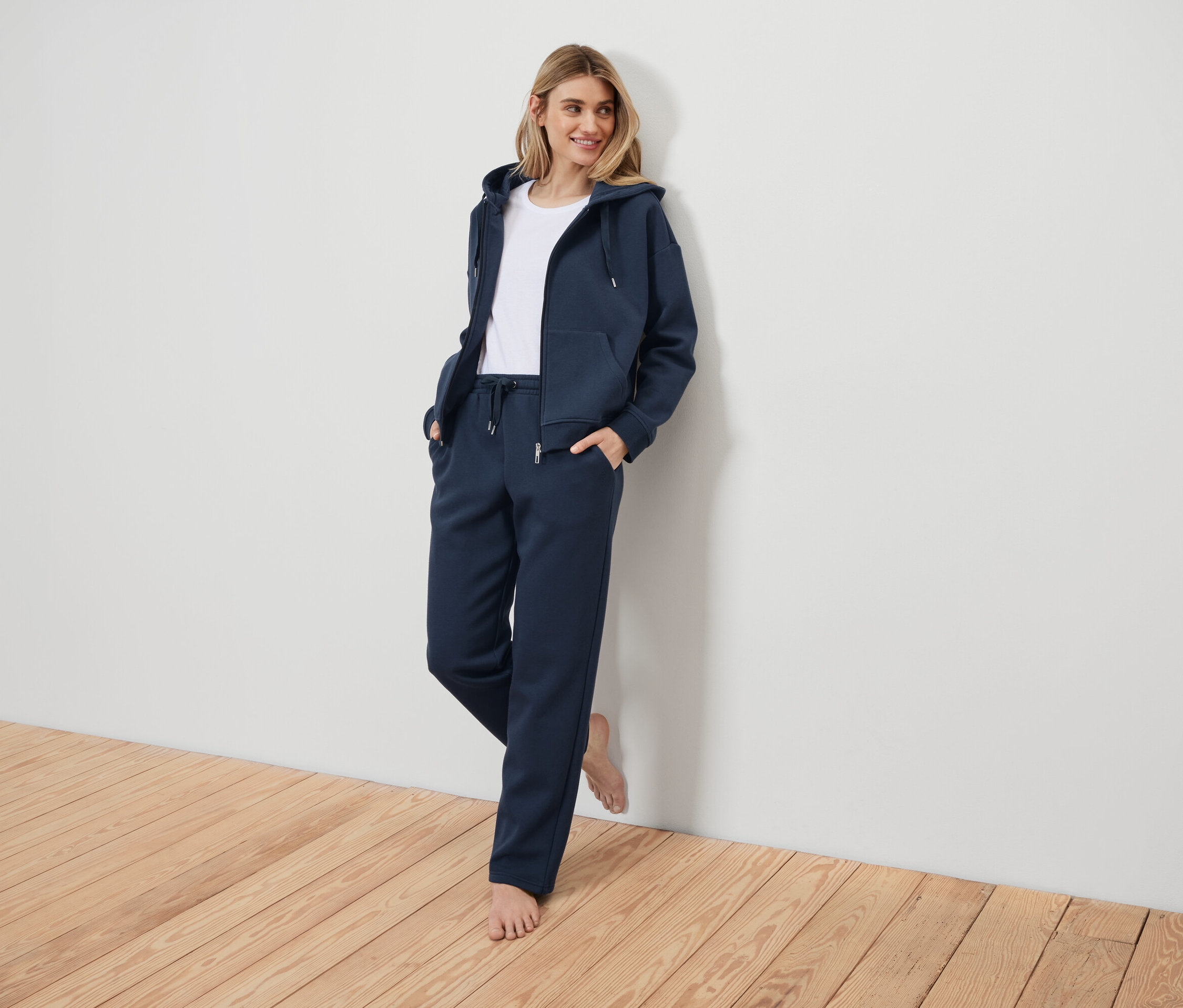 Kobieta w granatowym dresie oparta o białą ścianę.