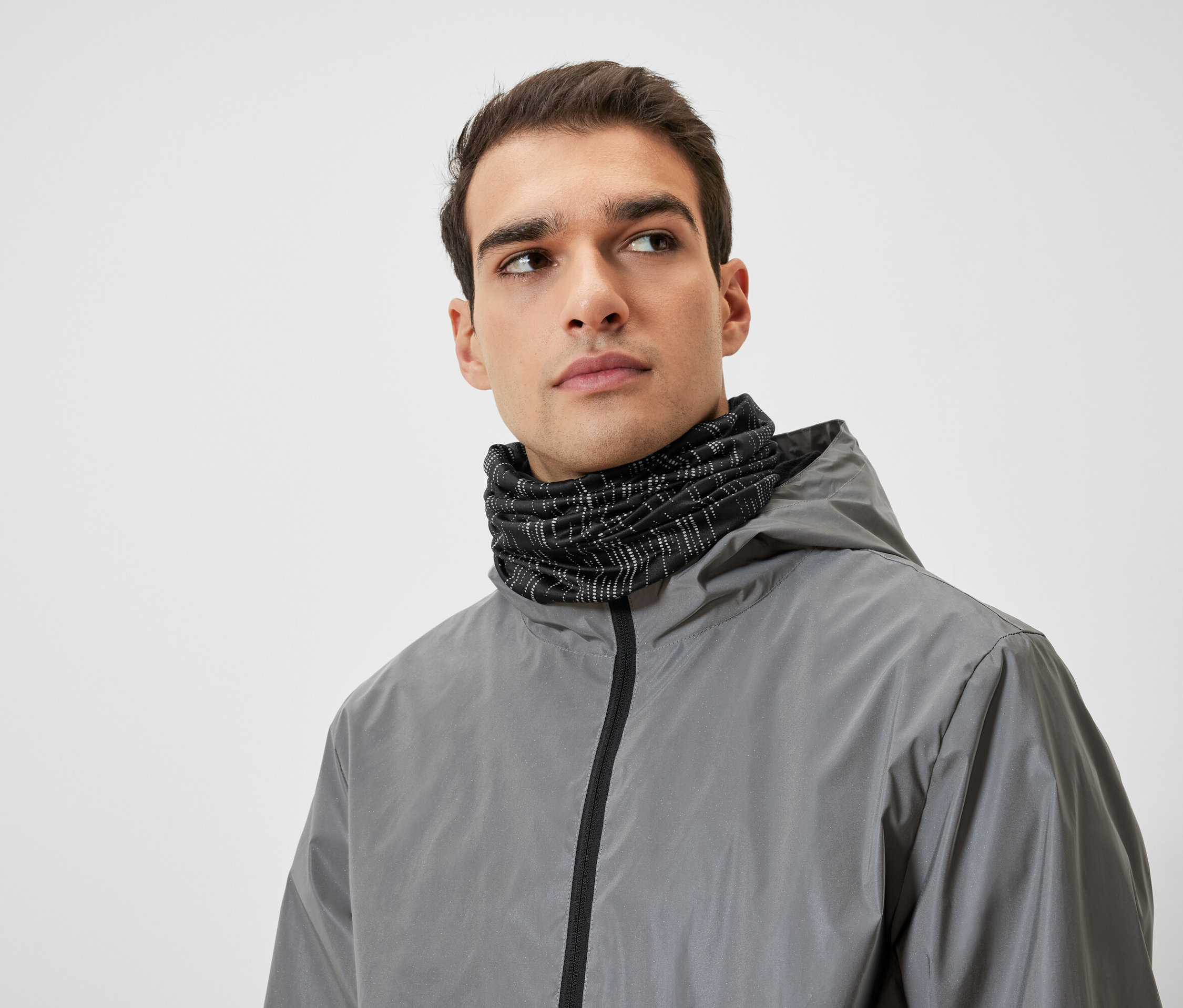 Mężczyzna o brązowych włosach ma na sobie szarą kurtkę z kapturem i czarny ocieplacz na szyję.