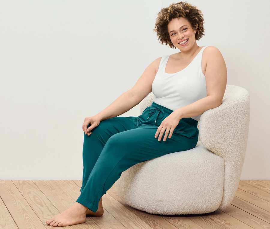 Kobieta siedzi w fotelu, ubrana w podkoszulki, 2 sztuki i spodnie od piżamy.