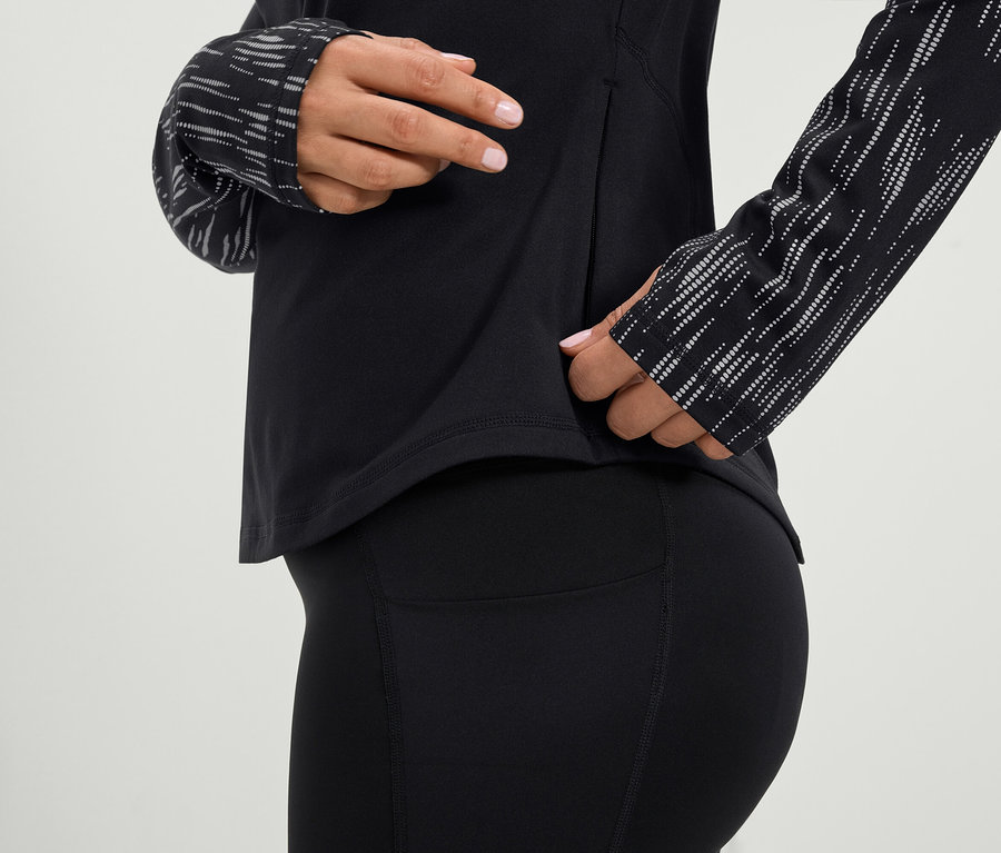 Kobieta sięga do bocznej kieszeni czarnej koszulki sportowej z długim rękawem i czarnych legginsów.