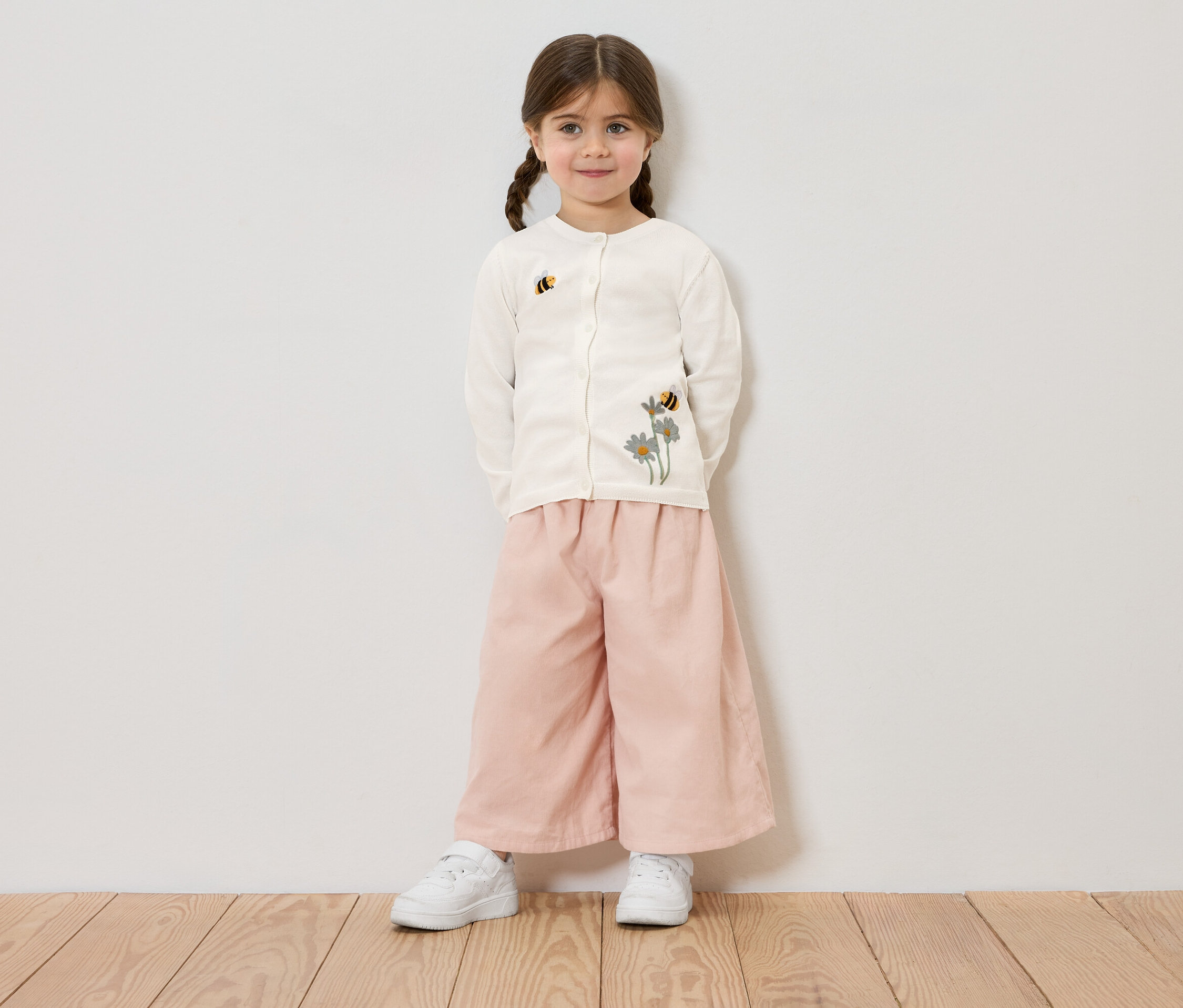 Dziewczynka z warkoczami ma na sobie kremowy kardigan z haftem kwiatowym i pszczół, różowe spodnie i białe trampki, na tle białej ściany.