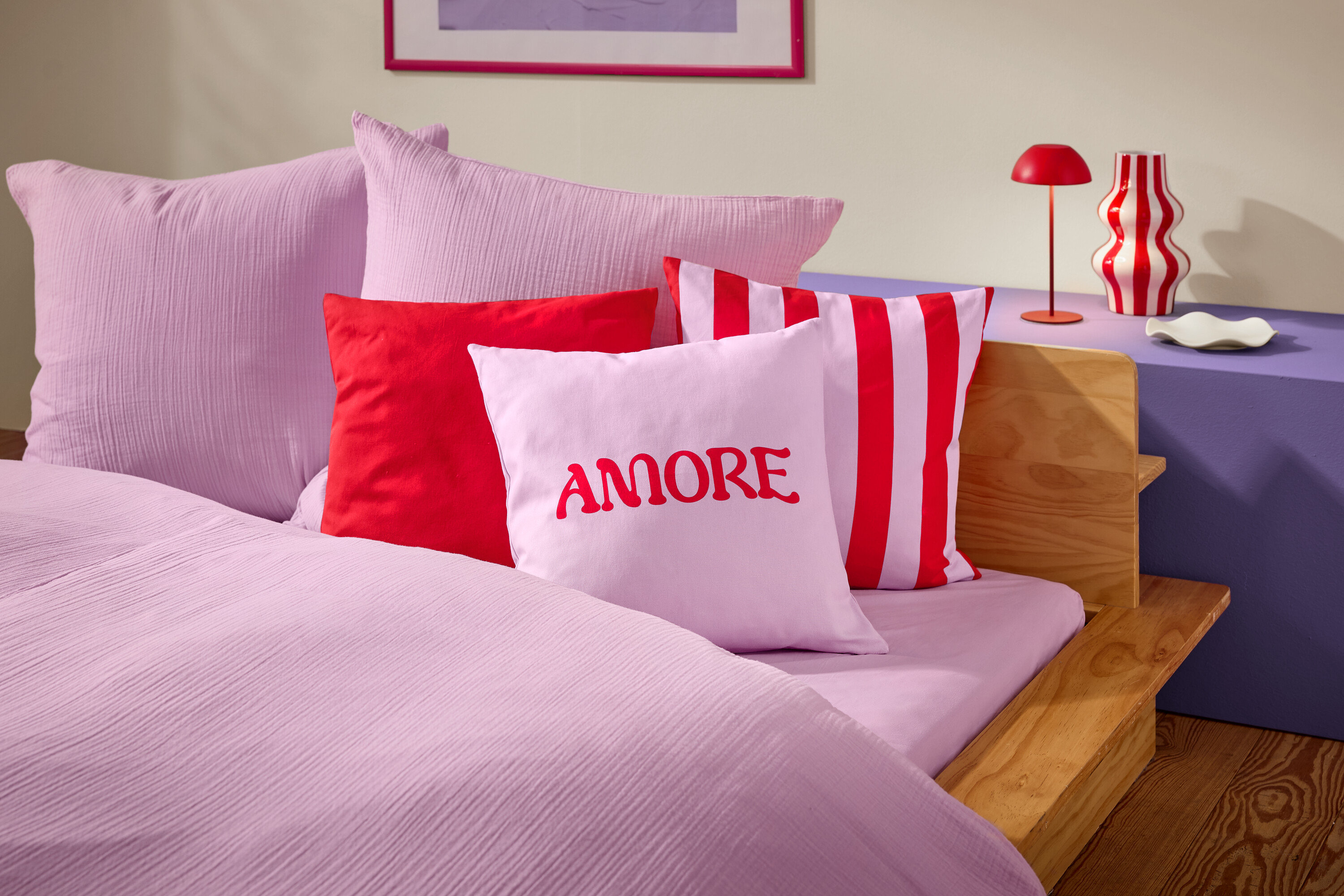 Zbliżenie łóżka z różową pościelą i poduszkami. Na jednej z poduszek widnieje słowo AMORE napisanym na czerwono.
