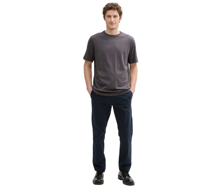 Mężczyzna o kręconych włosach stoi z rękami w kieszeniach spodni, ma na sobie szary T-shirt, niebieskie spodnie i czarne buty.