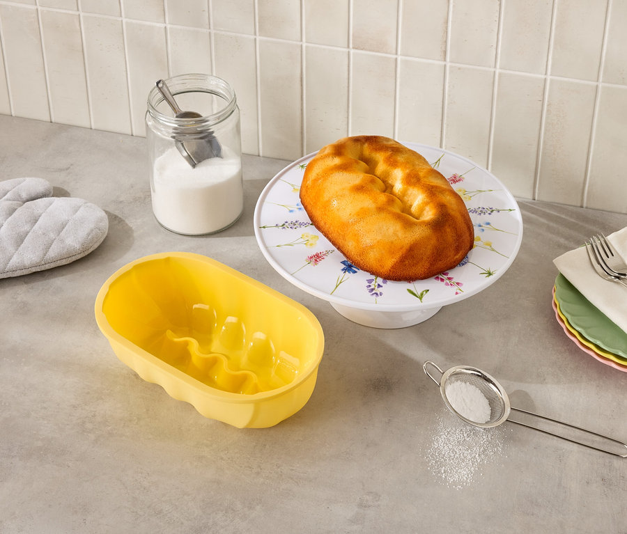 Na blacie kuchennym stoi babka na kwiatowym stojaku, żółta forma do babki, szklanka cukru, sito i łapki kuchenne.