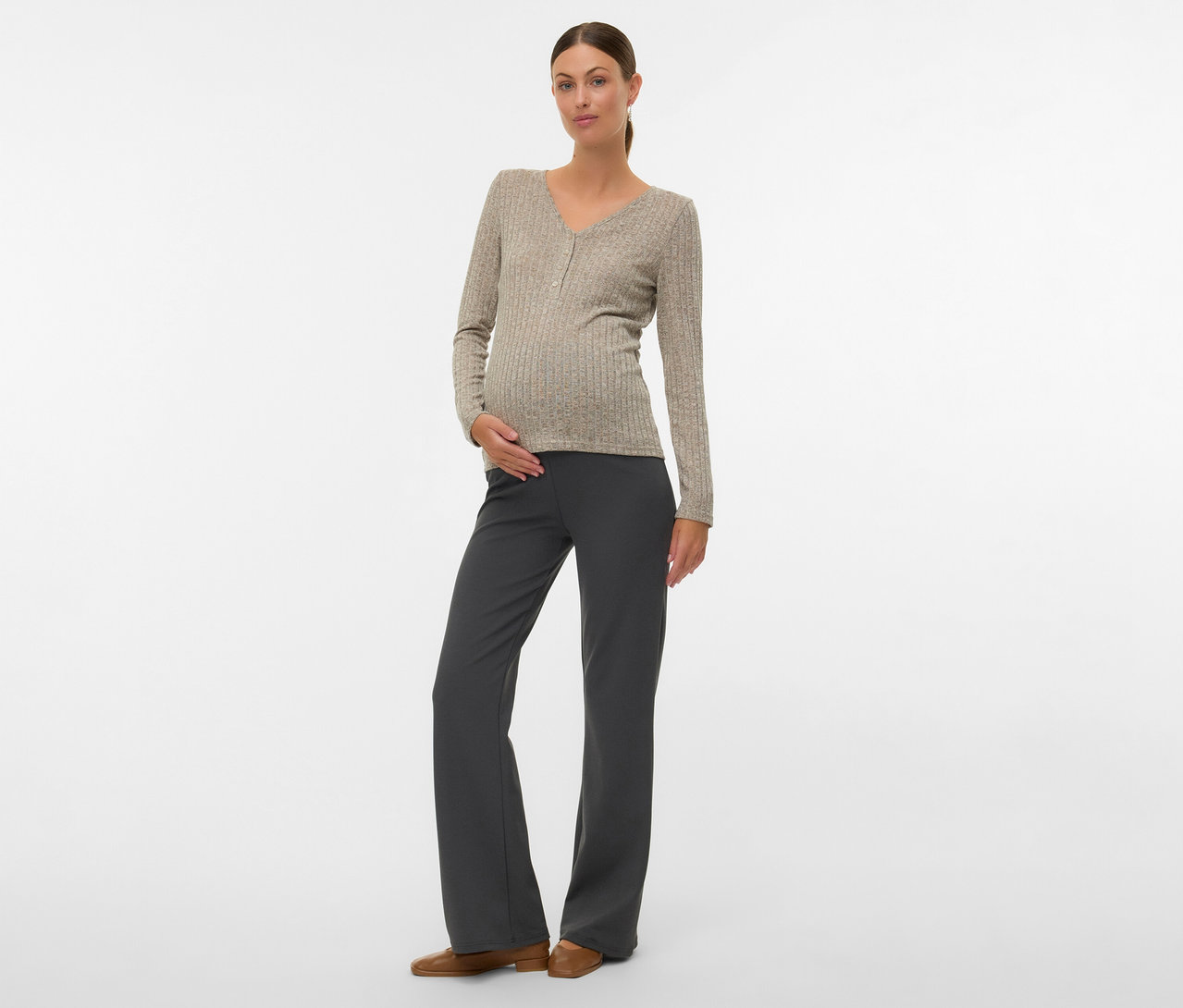 Kobieta z kokiem ma na sobie koszulkę MAMALICIOUS „MLPAMMIE LIA”, szare spodnie i brązowe buty, ręka na brzuchu.