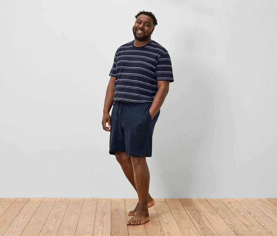 Uśmiechnięty mężczyzna stoi boso na drewnianej podłodze na tle białej ściany, ubrany w koszulkę w paski i granatowe szorty.