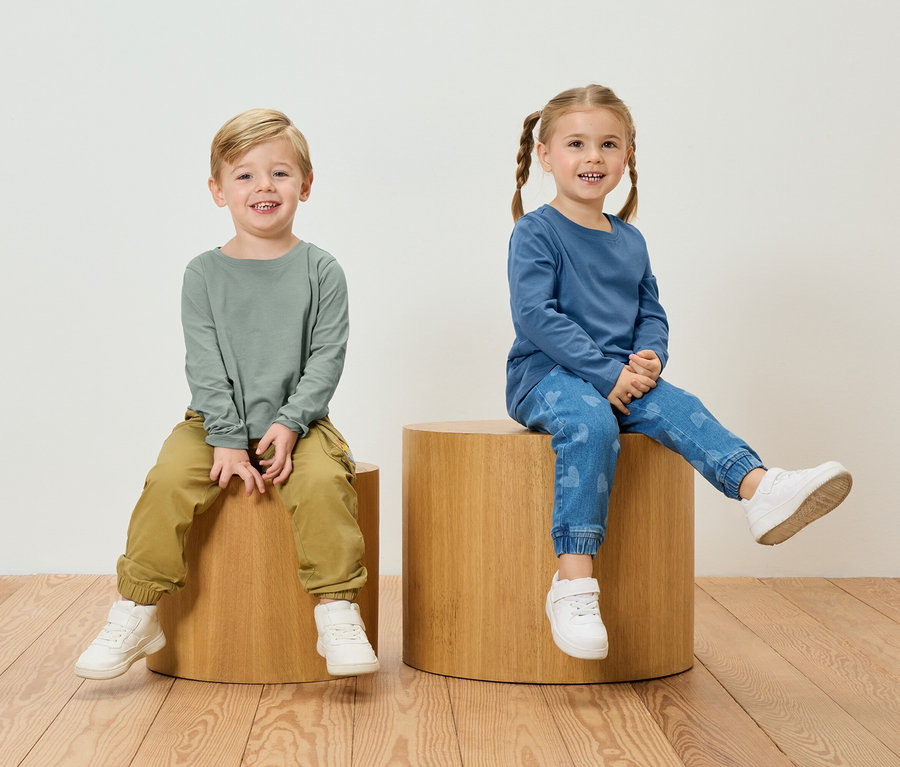 Chłopiec w zielonej koszulce z długim rękawem i spodniach khaki siedzi obok dziewczynki w niebieskiej koszulce z długim rękawem i dżinsach w serduszka na drewnianych stołkach.