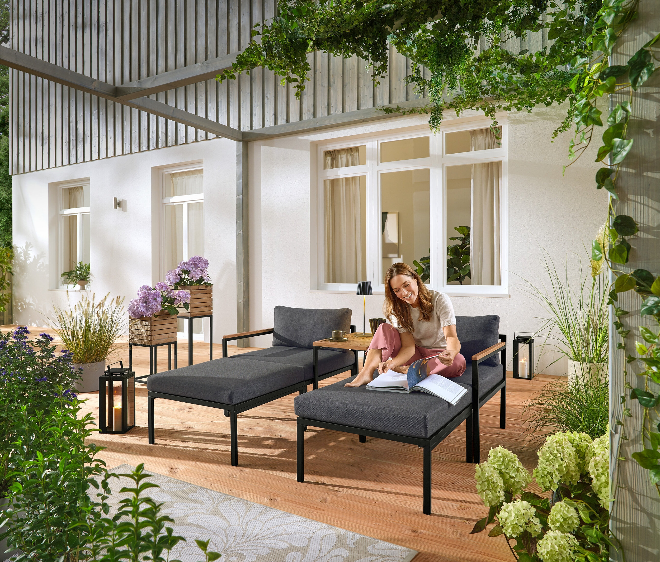 Kobieta siedzi na szarym leżaku ogrodowym i czyta książkę. W tle widoczne są dwie kolumny roślinne „Elin”.