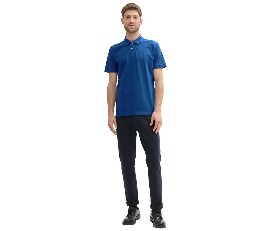 Mężczyzna w granatowych spodniach i niebieskiej koszulce polo Tom Tailor z dzianiny pique, Advanced Blue, z czerwonym logo, pozuje na białym tle.