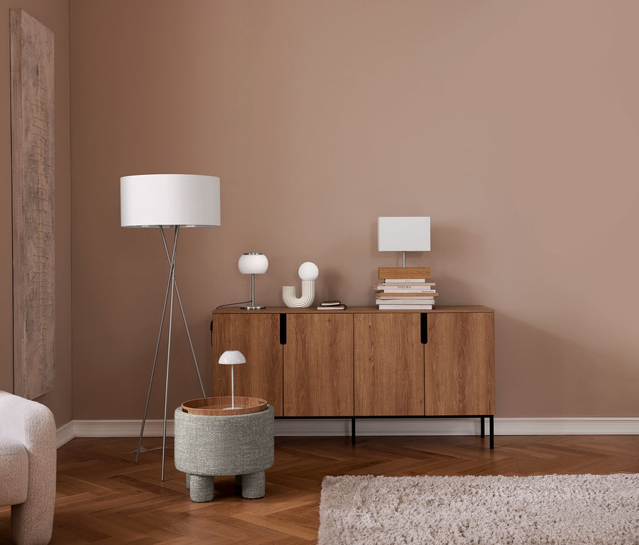 Nowoczesny salon z jasnobeżowym fotelem, drewnianą podłogą w jodełkę i ścianą w kolorze pudrowego różu. Srebrna lampa podłogowa na trójnogu stoi obok szarego, teksturowanego otomana z drewnianą tacą i małą białą lampką. Drewniana komoda mieści dwie białe lampy stołowe, przedmioty dekoracyjne i stosy książek. Na podłodze leży jasny, kudłaty dywan.