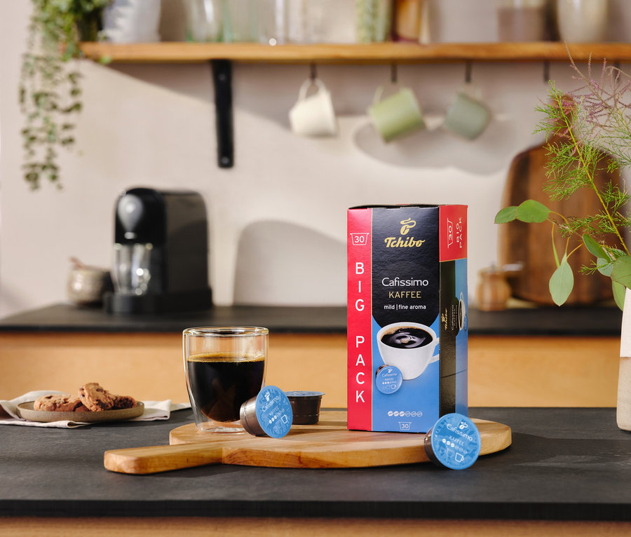 Na blacie stoi szklanka kawy obok opakowania Cafissimo pure+, black i dwóch kapsułek.