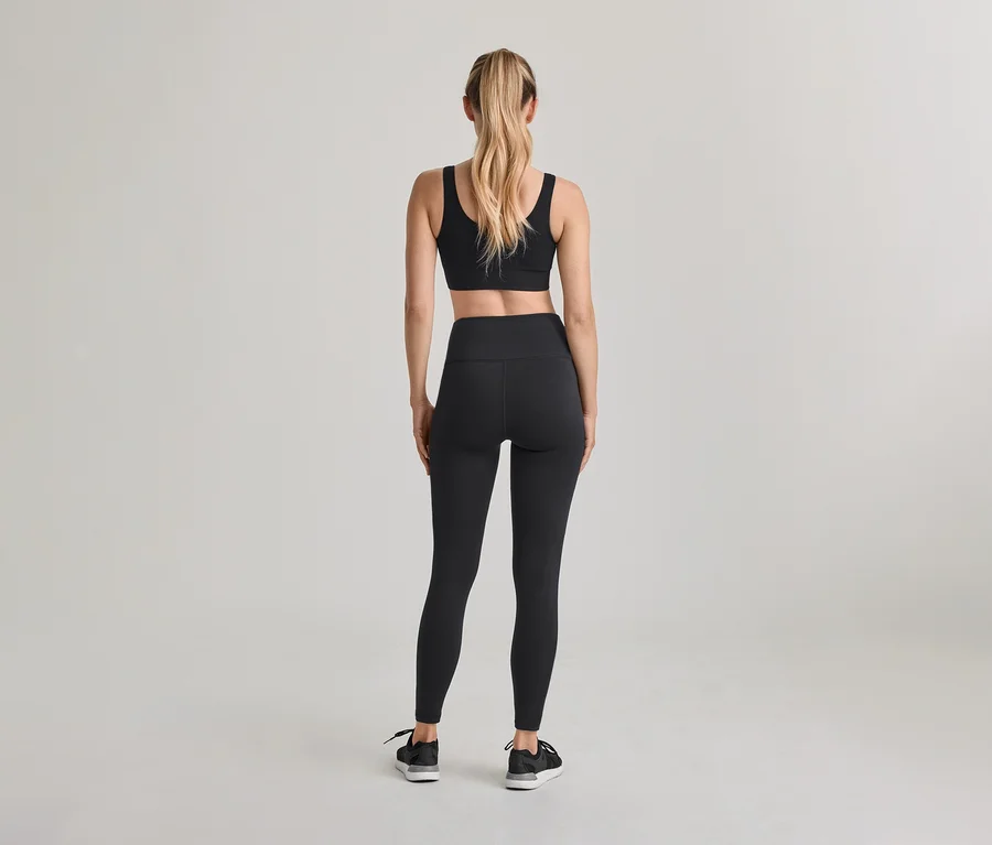 Kobieta stojąca tyłem, ubrana w czarny biustonosz sportowy, czarne legginsy z wysokim stanem i czarne sneakersy. Jej blond włosy są związane w kucyk.