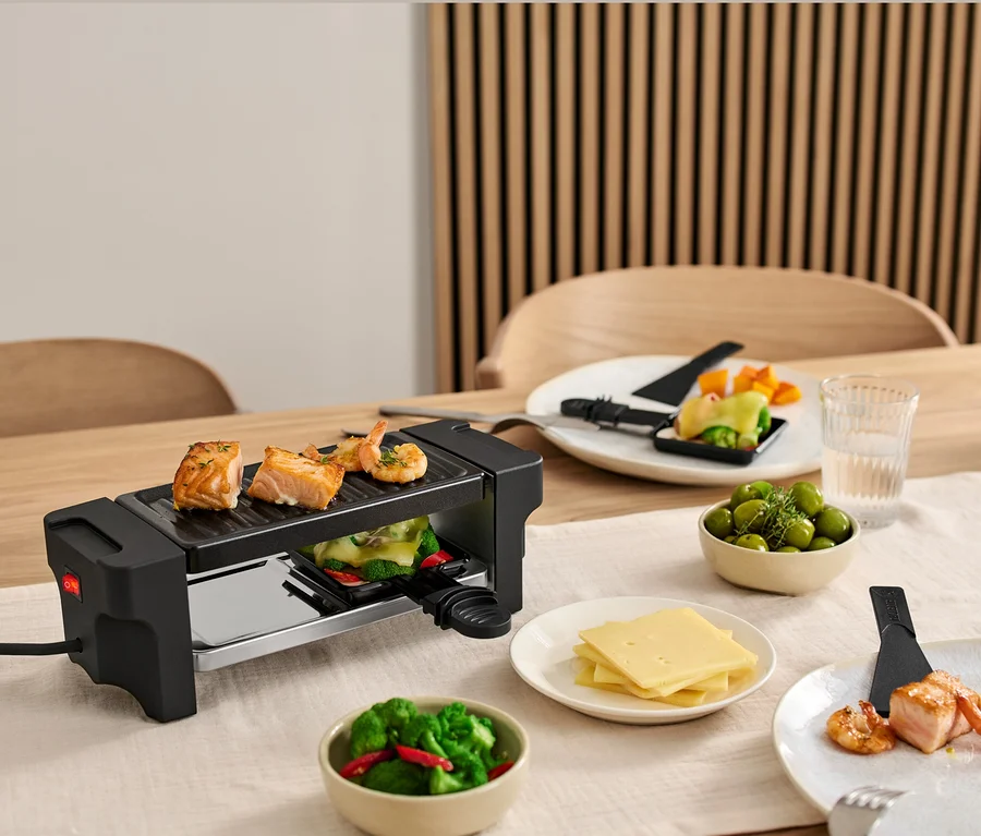 Raclette dla dwóch osób stoi na stole z łososiem i krewetkami na grillu. Talerze ze składnikami stoją wokół grilla.