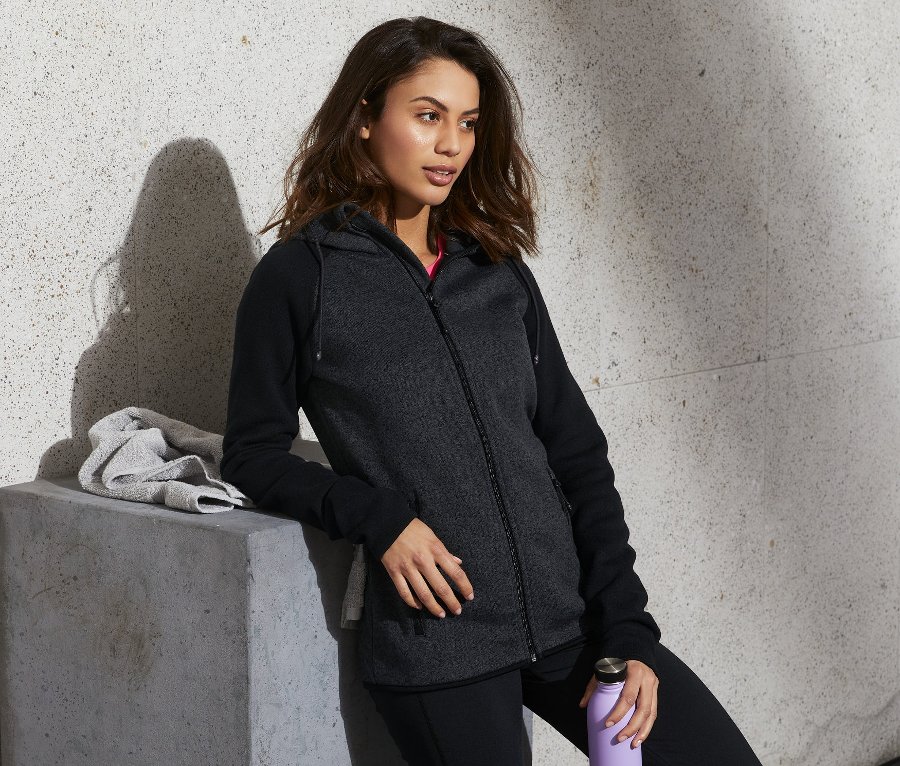 Kobieta opiera się o betonowy blok, ma na sobie rozpinaną bluzę z dzianiny polarowej, czarne spodnie i trzyma fioletową butelkę z wodą.