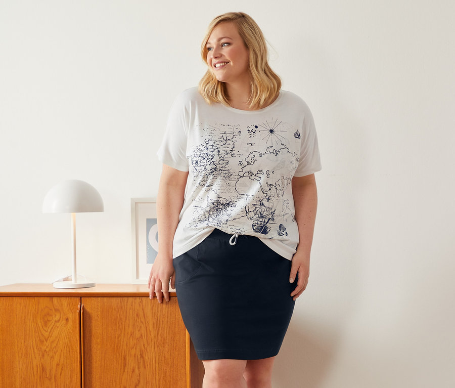 Kobieta stoi obok szafki, ubrana w białą bluzkę z okrągłym wycięciem pod szyją i spódnicę dresową.