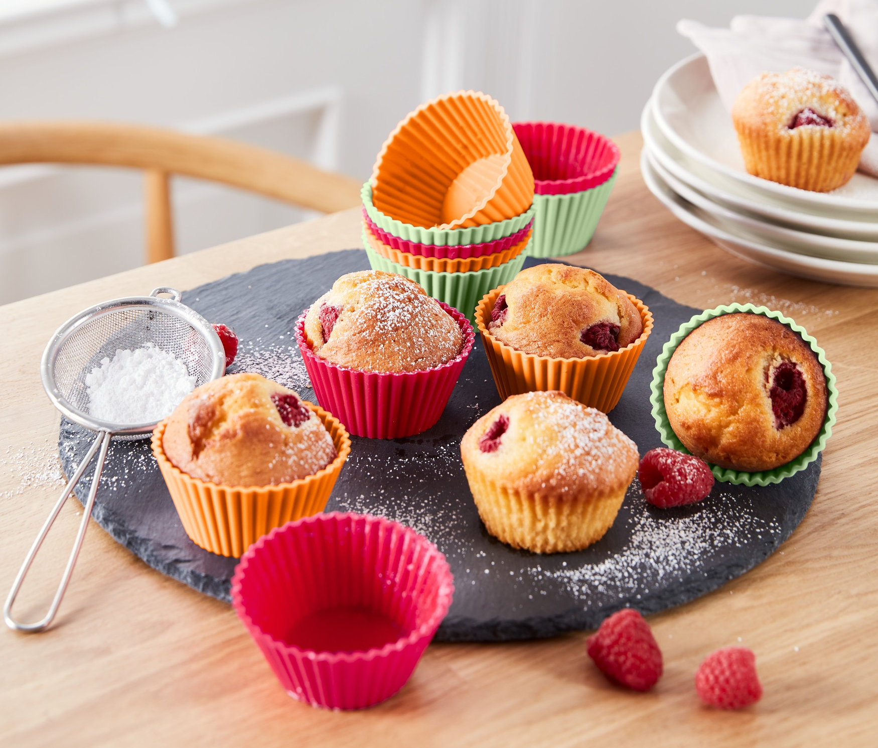 Muffiny w kolorowych foremkach do muffinek, 12 sztuk, posypane cukrem pudrem leżą na stole.