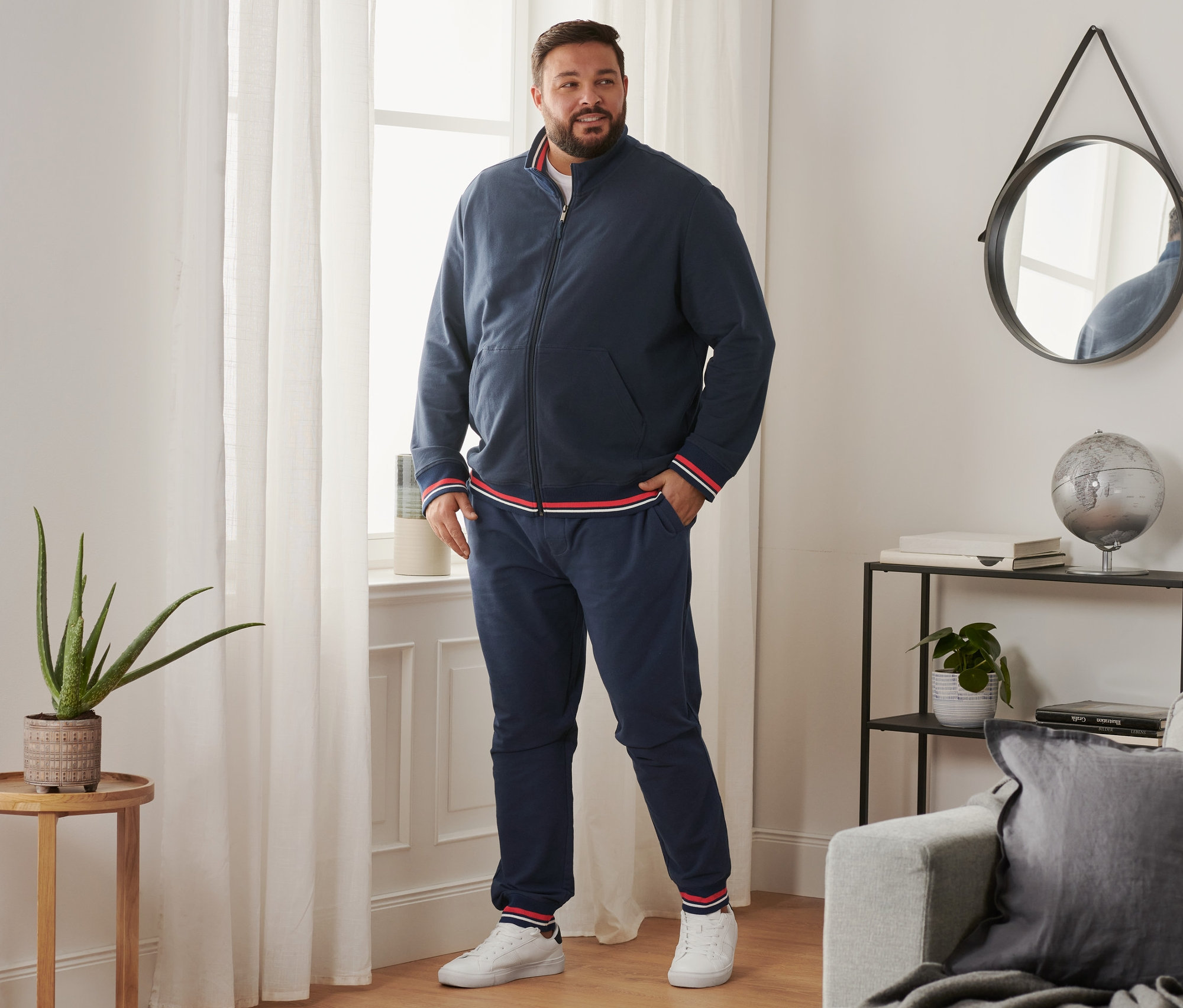 Mężczyzna stoi przy oknie, ubrany w spodnie dresowe, buty typu sneaker i bluzę z dzianiny dresowej.