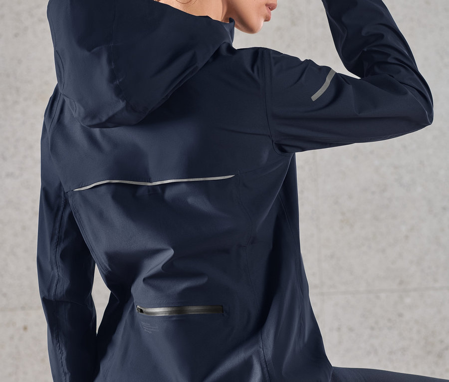 Kobieta ociera pot z czoła, ma na sobie granatową kurtkę przeciwdeszczową do biegania.