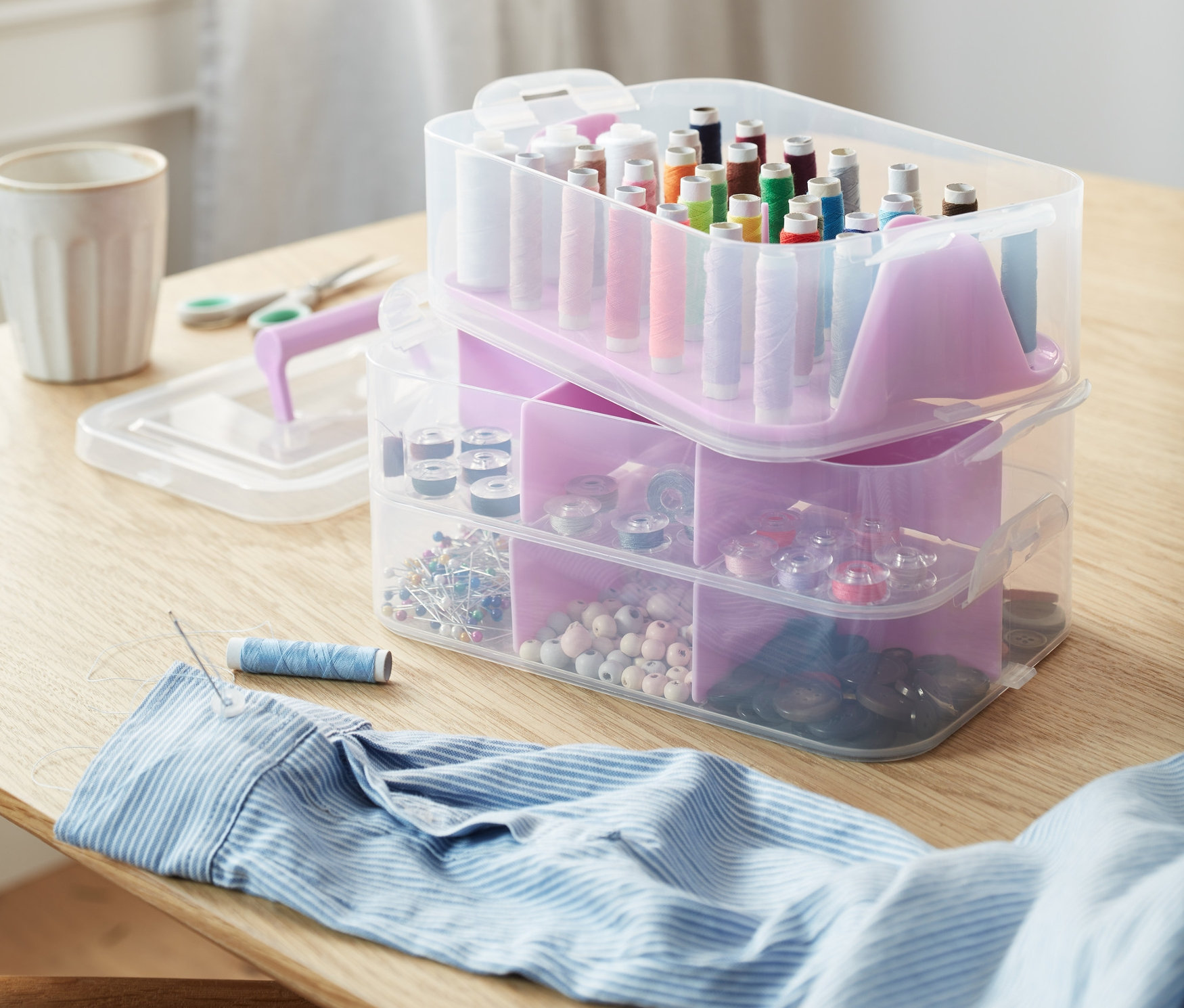 Na stole leżą pudełka do przechowywania, nożyczki, guziki, szpulki nici i igła z nitką w koszuli.