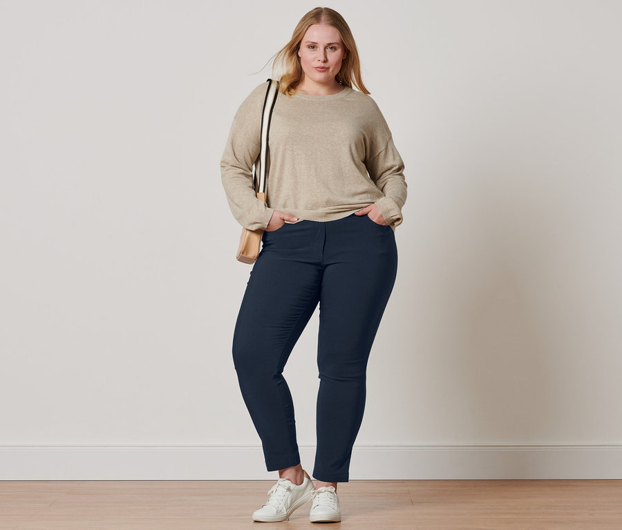 Kobieta pozuje w beżowym swetrze z delikatnej dzianiny ze ściągaczem, granatowych spodniach z bengaliny, skórzanych butach typu sneaker i z torbą na ramię.
