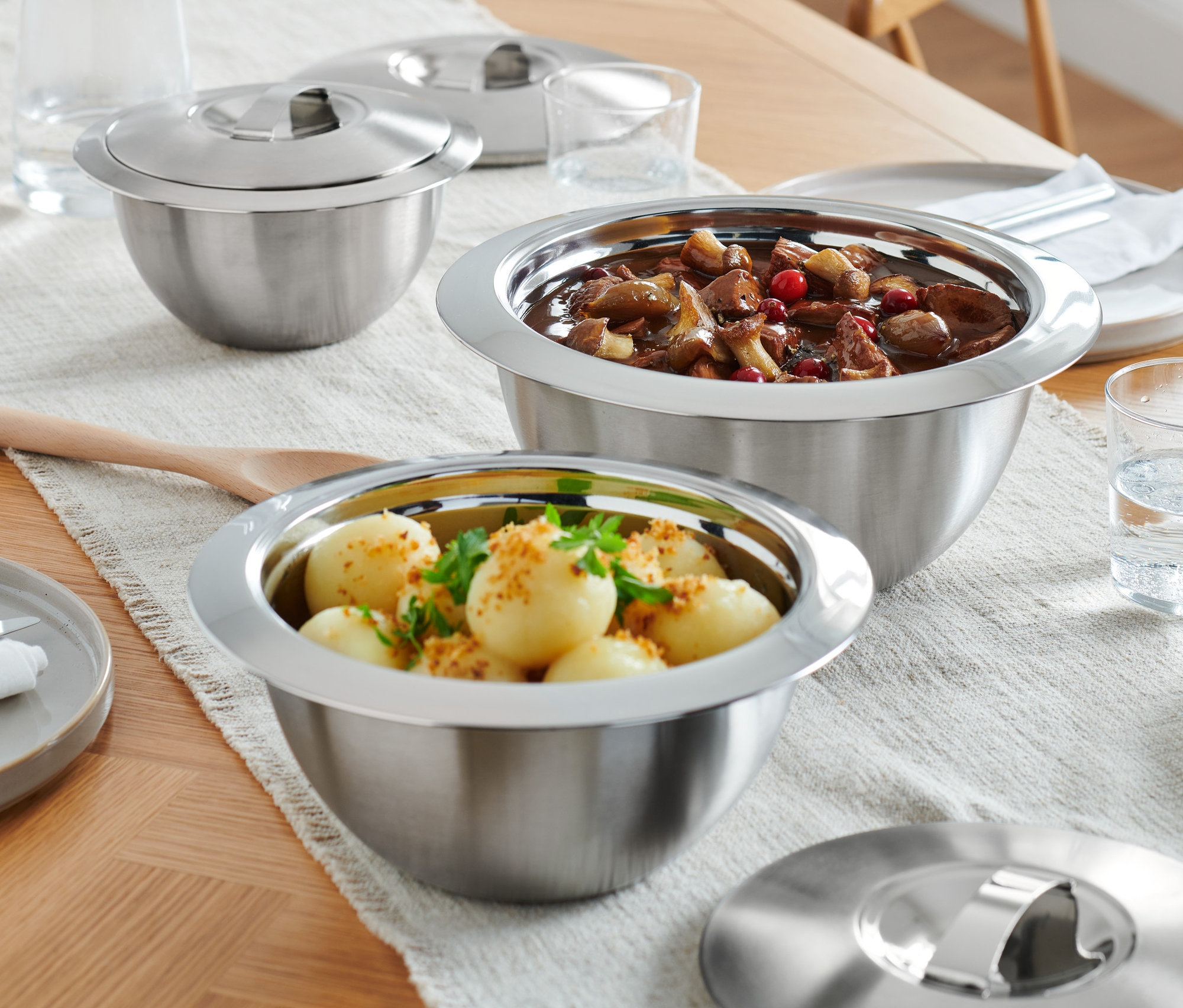 Na drewnianym stole stoją trzy miski do serwowania utrzymujące temperaturę z pokrywkami, talerze i szklanki z wodą.