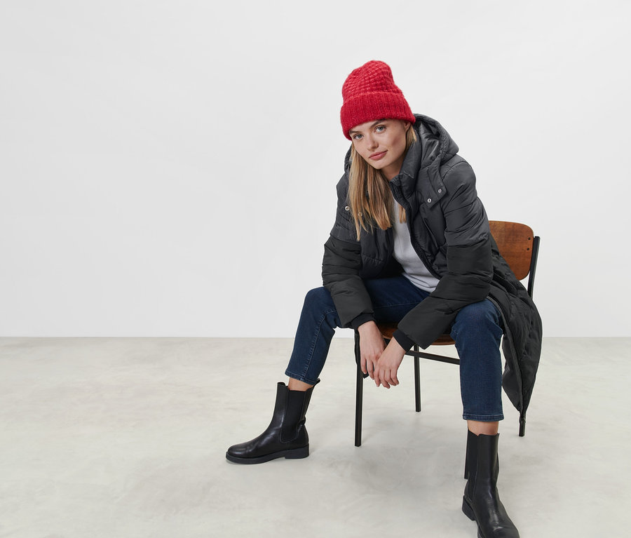 Kobieta siedzi na krześle, ubrana w płaszcz pikowany z kapturem i czerwoną czapkę.