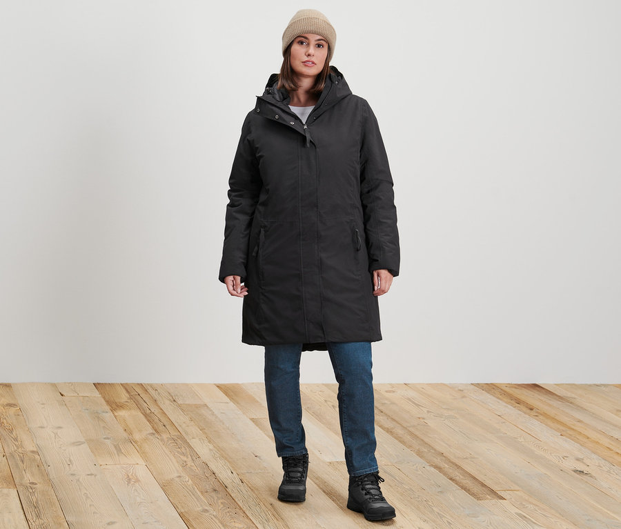 Kobieta stoi w czarnym płaszczu ciepłochronnym, ciemnoniebieskich jeansach slim »Fit Emma« i piaskowej czapce z dzianiny.