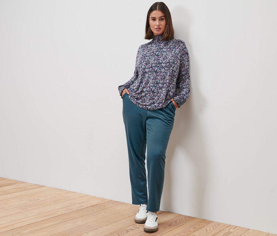 Kobieta pozuje w bluzce ze stójką we wzór millefleurs, spodniach dresowych i skórzanych sneakersach.