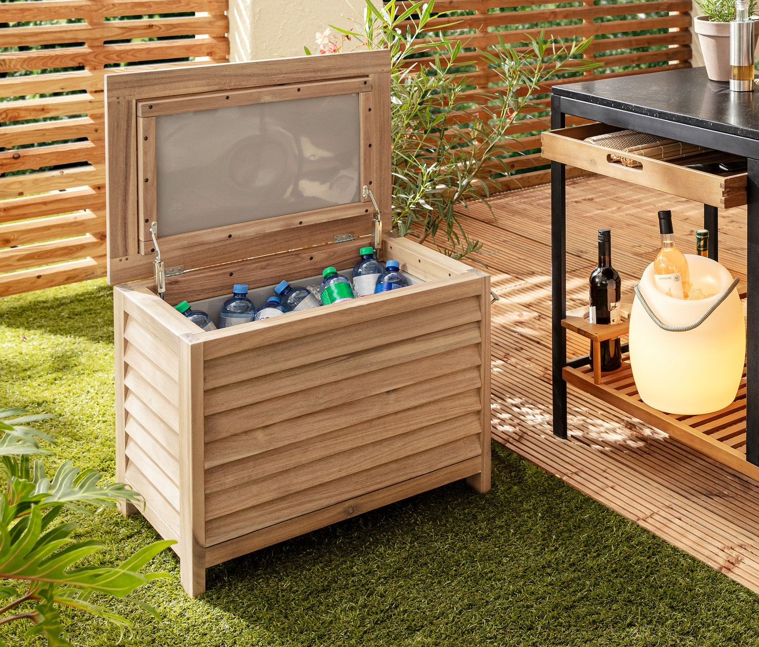 Na trawniku stoi dekoracyjna lodówka turystyczna z otwieraczem do butelek, obok niej wózek barowy „Elin” z szufladą, na stole akumulatorowa lampa stołowa LED i schładzacz do butelek z oświetleniem LED.