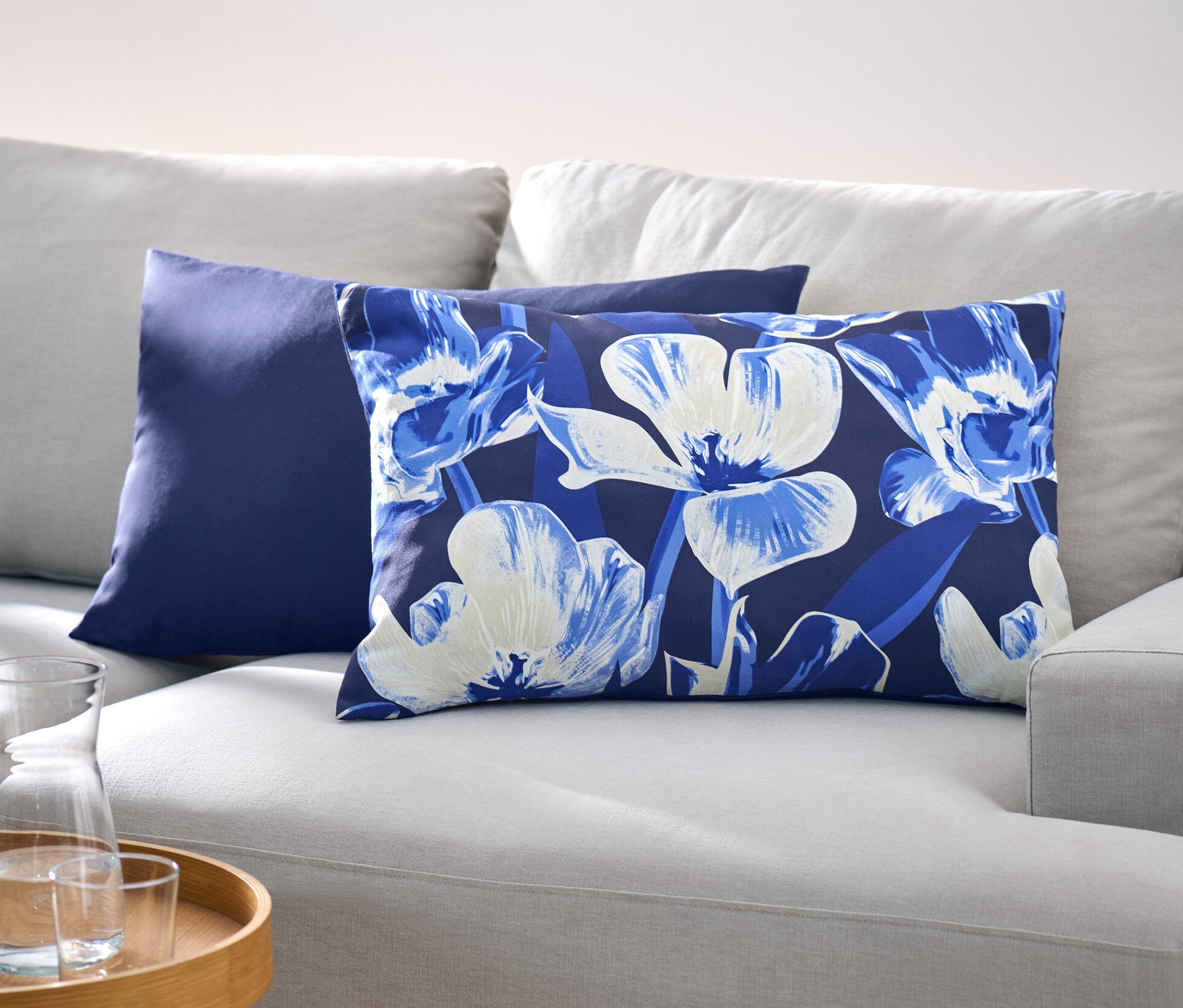 Na jasno szarej sofie leżą dwie dekoracyjne poszewki na poduszki, jedna jest ciemnoniebieska, a druga ma kwiatowy wzór.