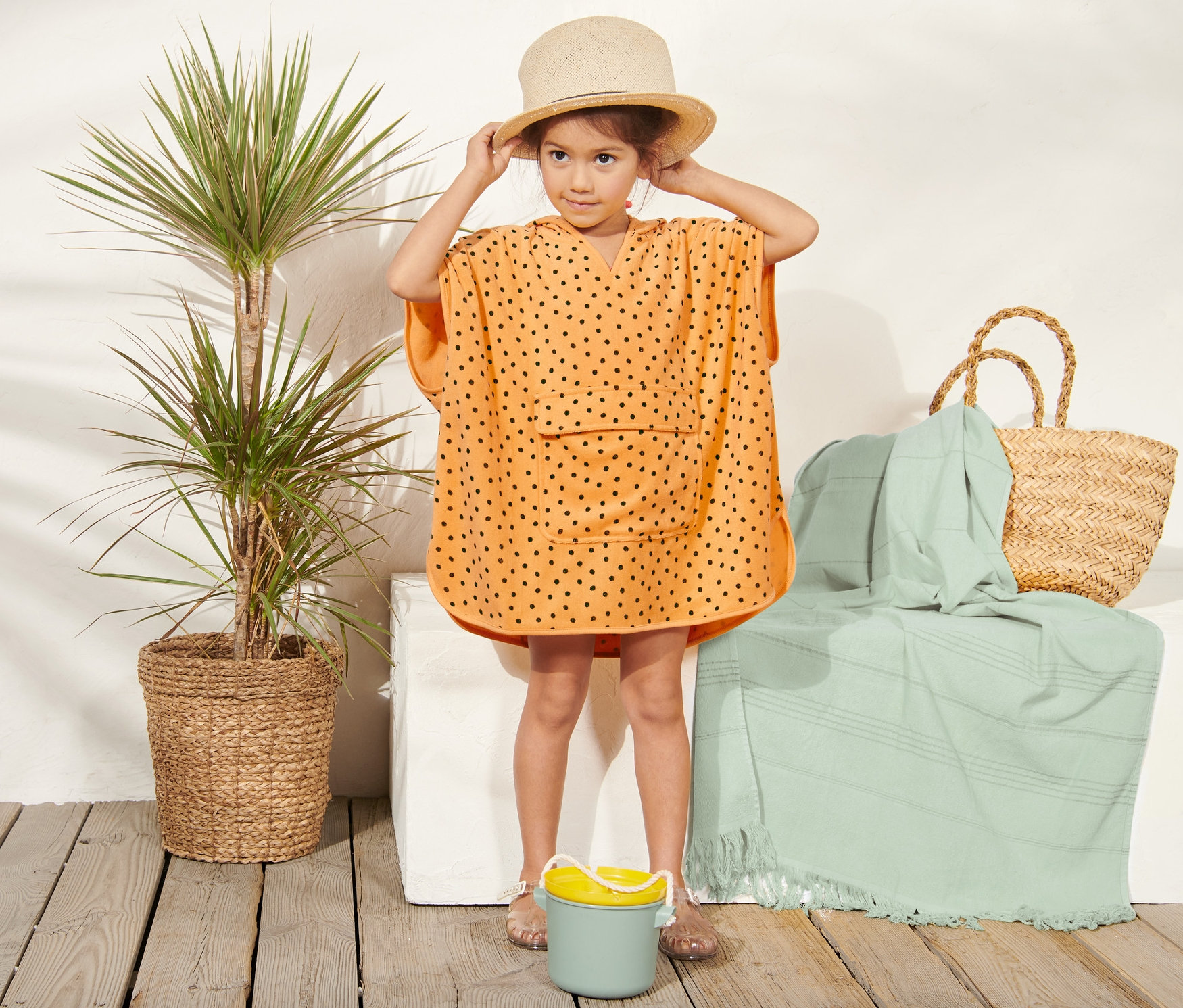 Dziewczynka w pomarańczowym poncho frotte w czarne kropki i słomkowym kapeluszu stoi obok rośliny w koszyku i torby plażowej.
