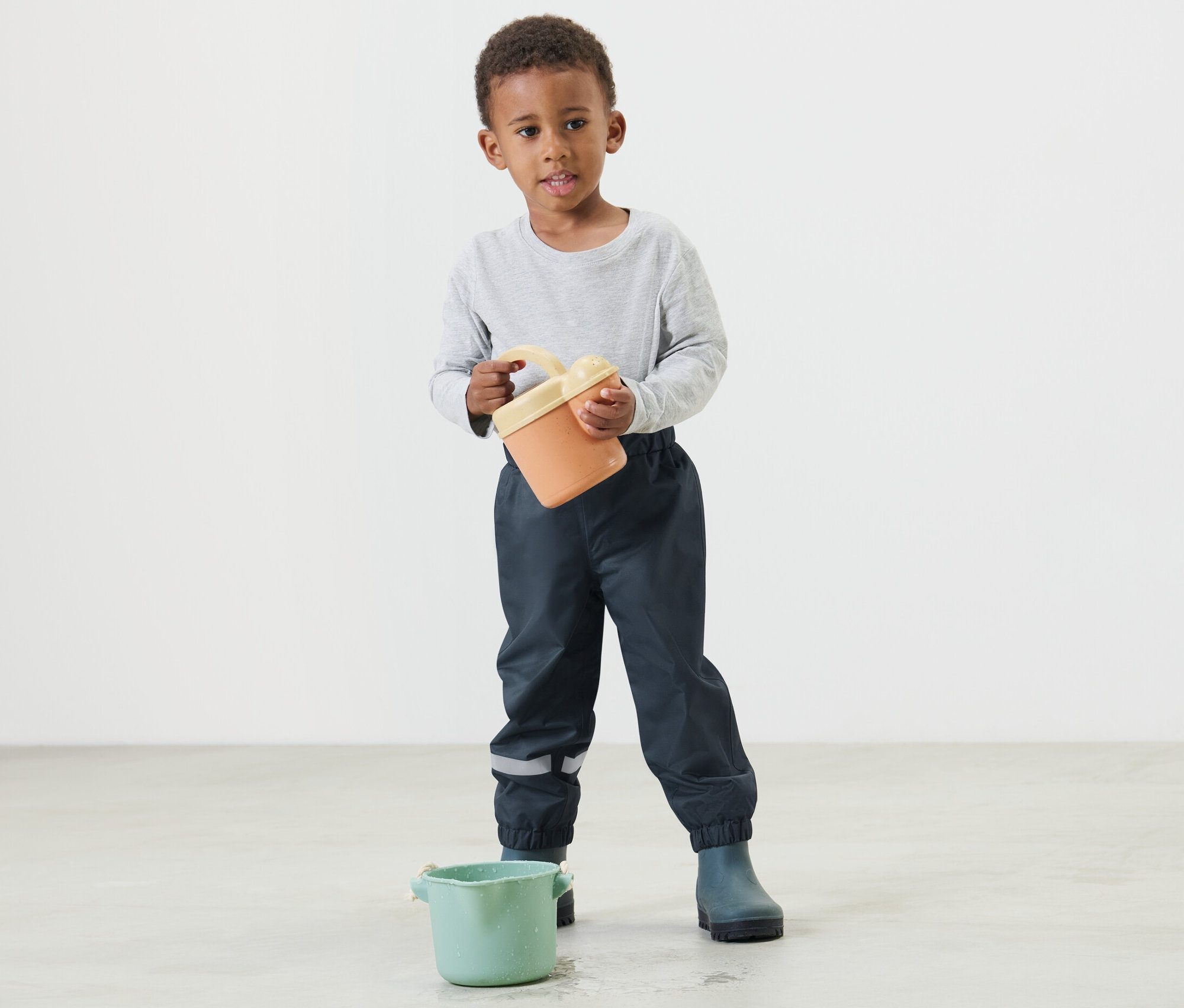 Chłopiec trzyma pomarańczowe wiadro i ma na sobie granatowe spodnie przeciwdeszczowe.