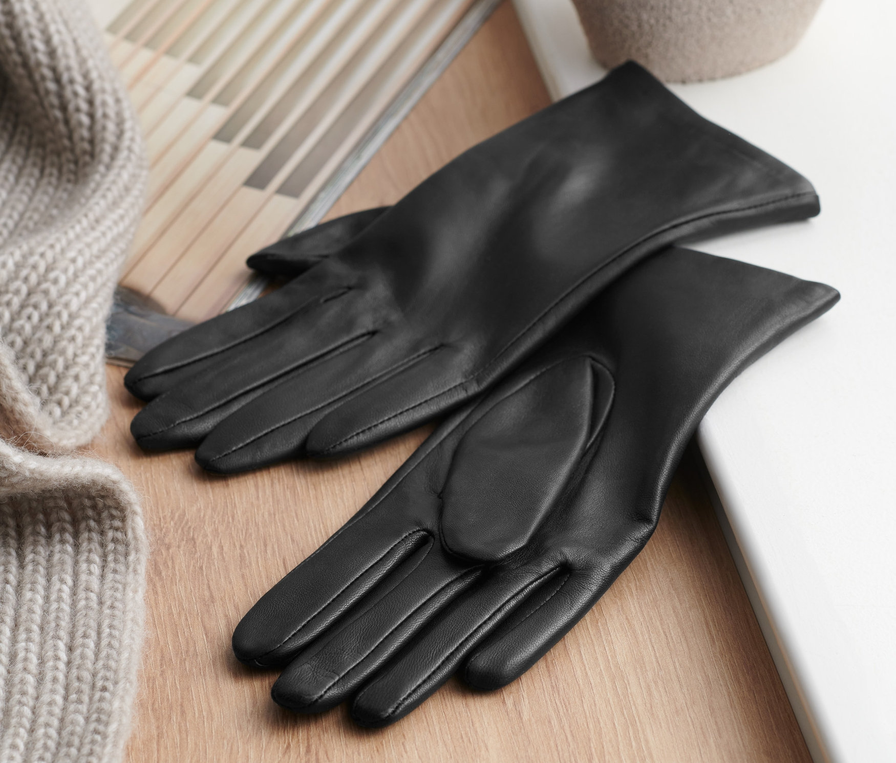 Czarne skórzane rękawiczki leżą na drewnianym stole obok swetra i wazonu.